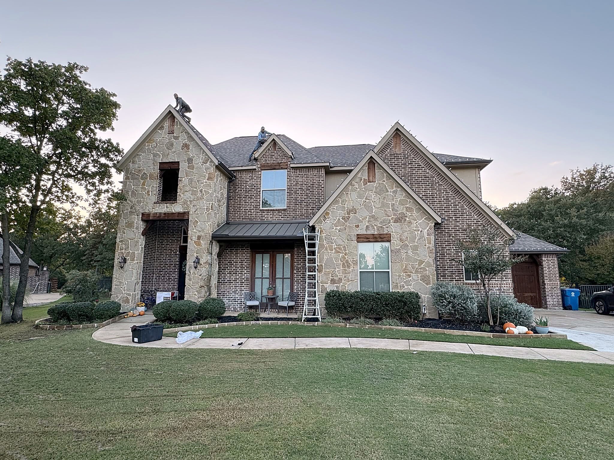 A two-story stone and brick home at dusk with a ladder against the roofline and two roofers on the ridge